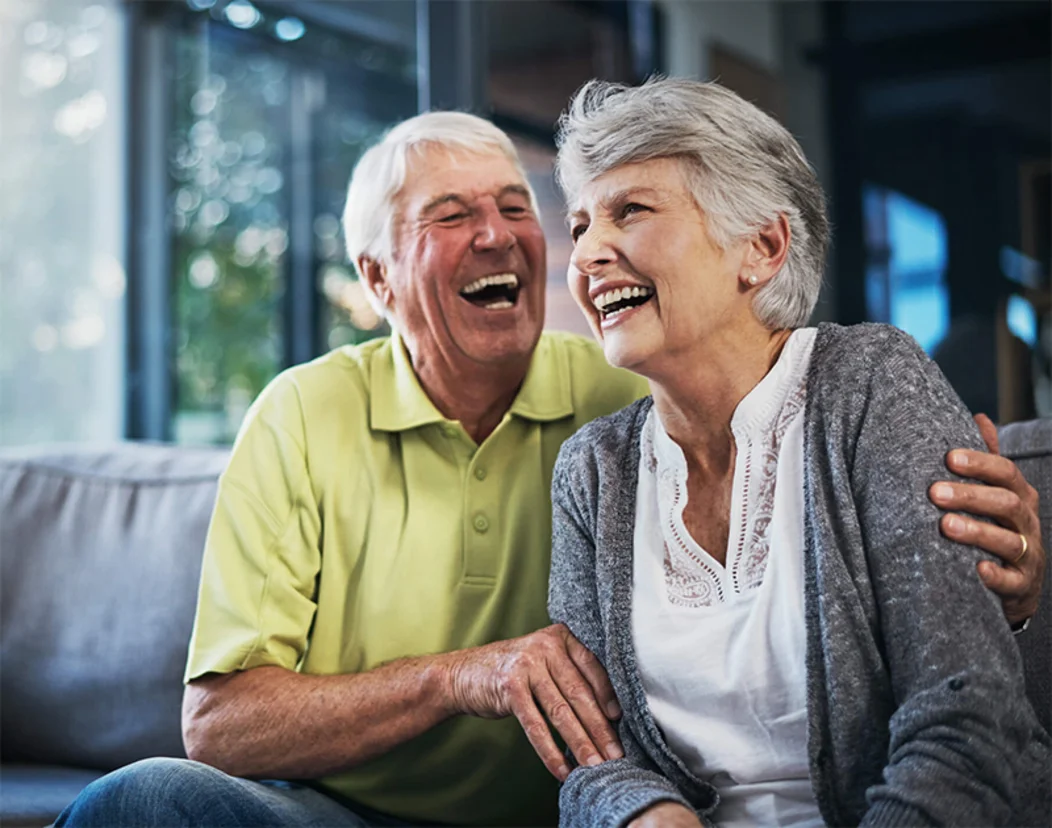 Casal de idosos sorrindo, sentados no sofá de casa